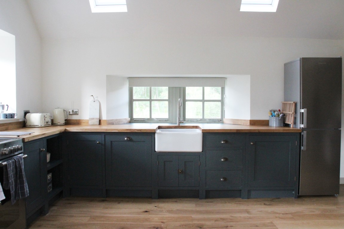 A modern kitchen in Pheasant Cottage features dark cabinets, a white farmhouse sink, wooden countertops, and a stainless steel fridge by a window.