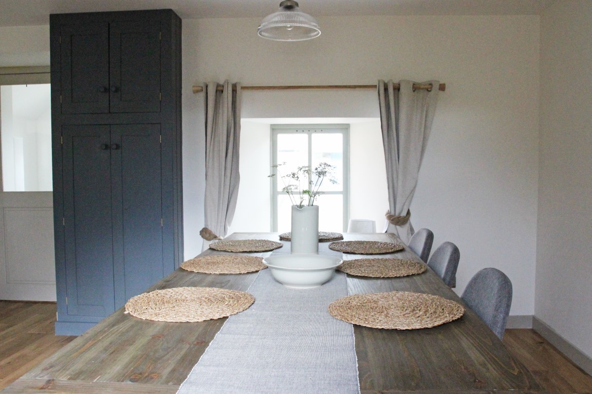 In the charming Pheasant Cottage dining room, a long wooden table surrounded by six chairs awaits, adorned with woven placemats and a vase brimming with flowers, all elegantly illuminated by the ceiling light.