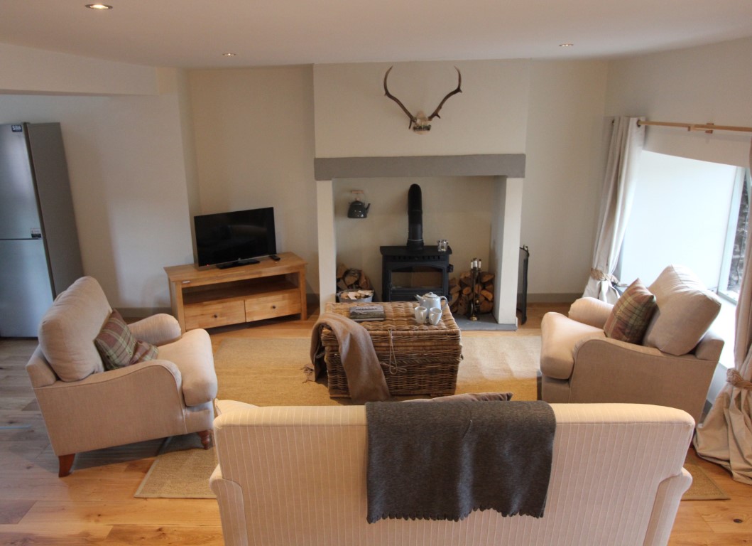 A cozy cottage living room features beige sofas, a TV, and a welcoming fireplace. A woven coffee table complements the rustic charm, while antlers mounted above the mantel add character alongside tasteful pheasant accents.