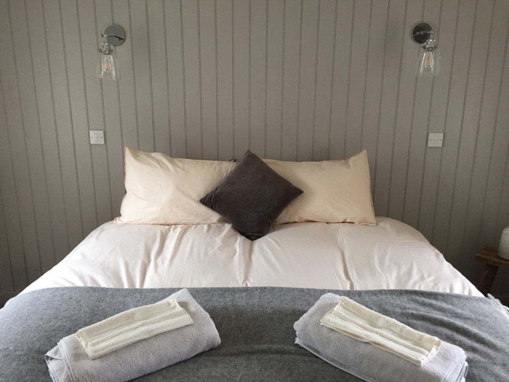 A neatly made bed with beige and gray bedding, topped with three pillows and two folded towels at the foot, under wall lights in charming Pheasant Cottage.