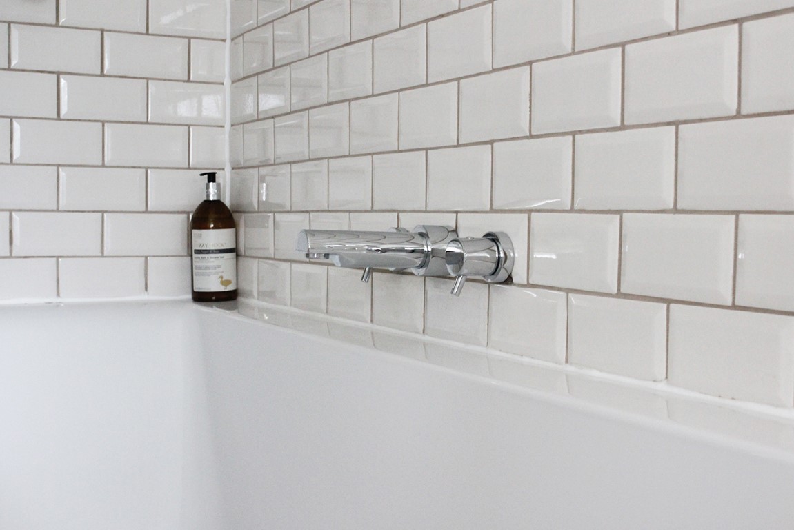 The minimalist bathtub at Pheasant Cottage features a glossy white tile wall, chrome faucet, and a brown bottle of soap perched on the edge.