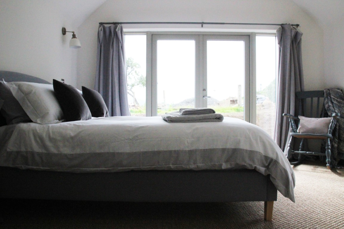 A neatly made bed with gray and white linens sits in Pheasant Cottage, next to large windows with curtains, complemented by a wooden chair in this simple bedroom.