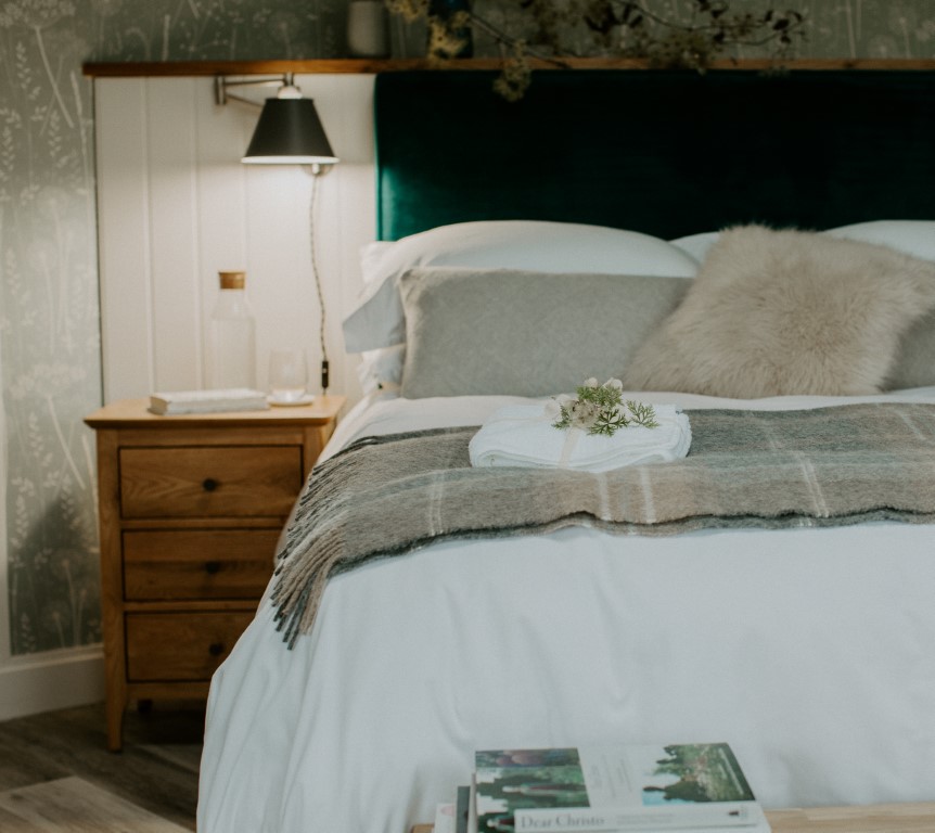 Cozy bedroom retreat with a neatly made bed, decorative pillows, a nightstand holding a lamp and water bottle, and books on a nearby table.