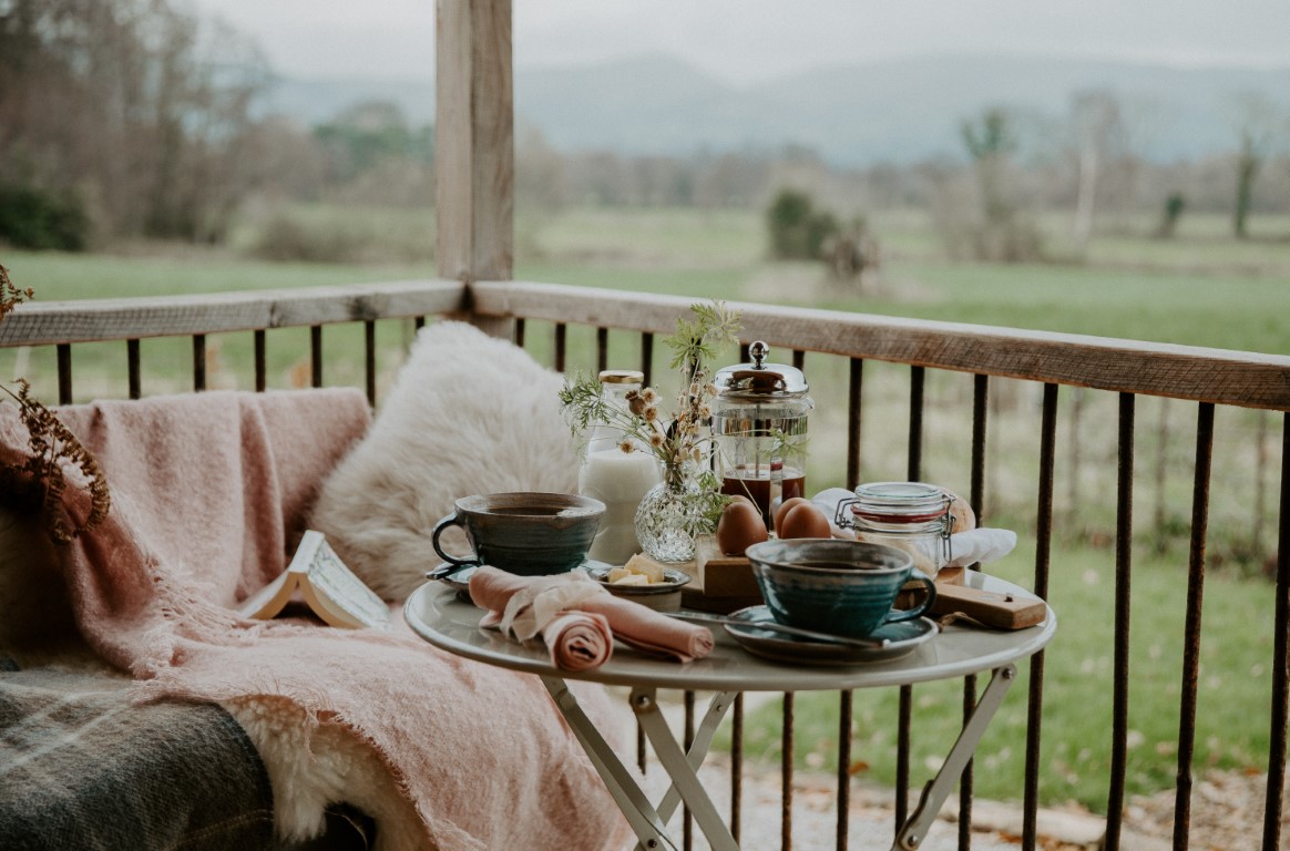 Cozy outdoor seating with a small table, two cups, a French press, and breakfast items on a porch overlooking a green landscape offers the perfect morning retreat.