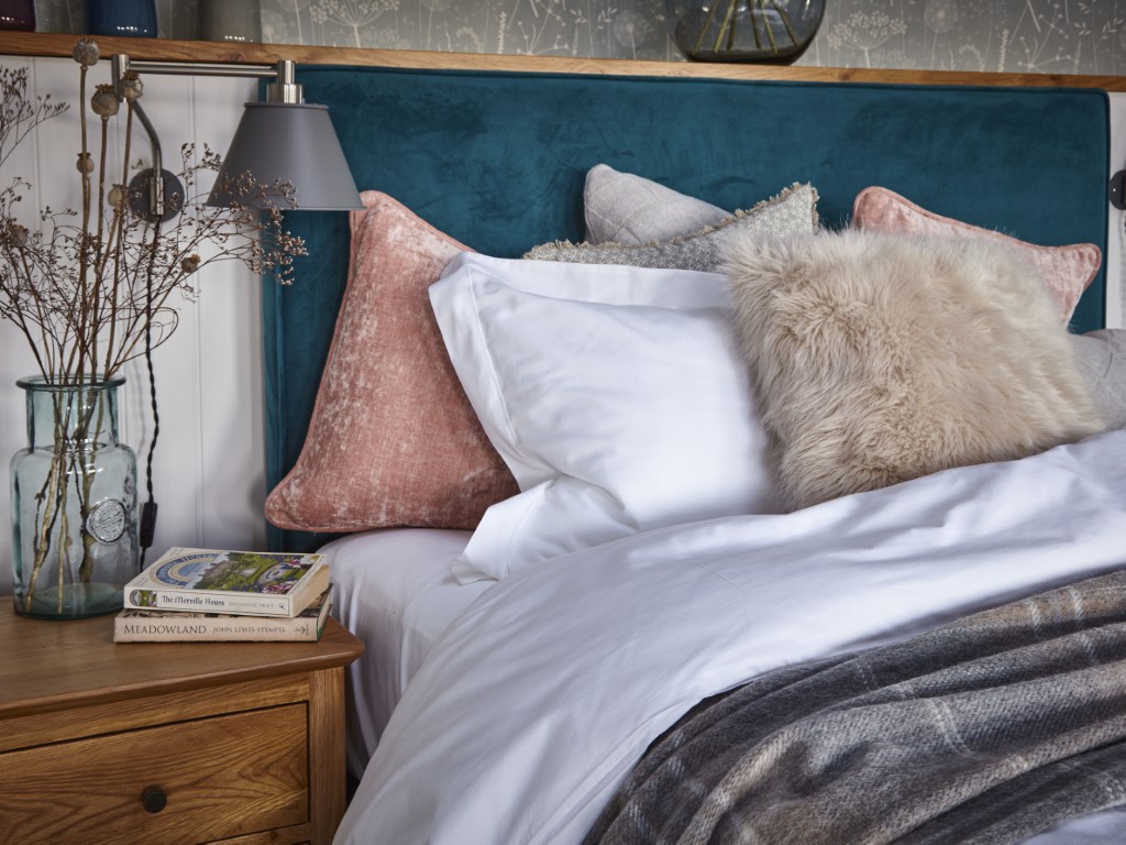 Cozy bedroom with a teal headboard, fluffy pillows, and a wooden nightstand adorned with books and a glass vase of dried flowers—a perfect retreat for relaxation after enjoying our convenient laundry service.