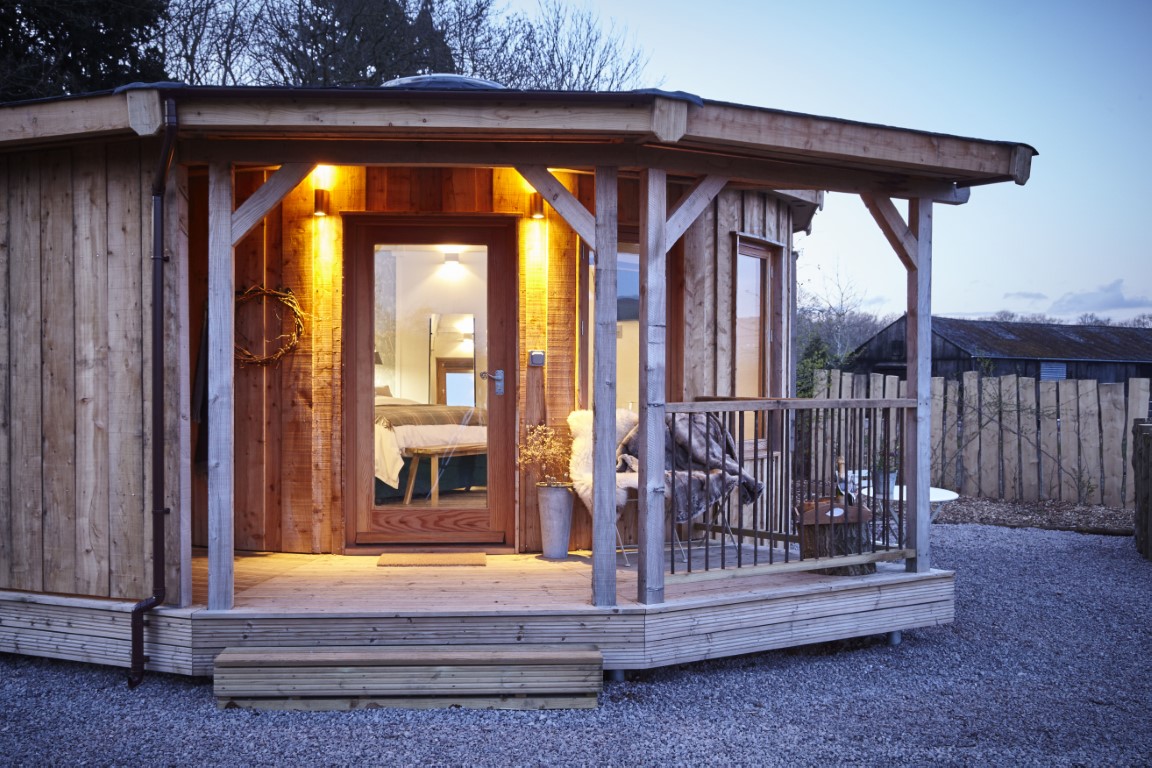 A round wooden cabin with a porch is warmly lit at dusk, offering a perfect retreat. A glimpse of a bed through the door invites relaxation after the day's end.