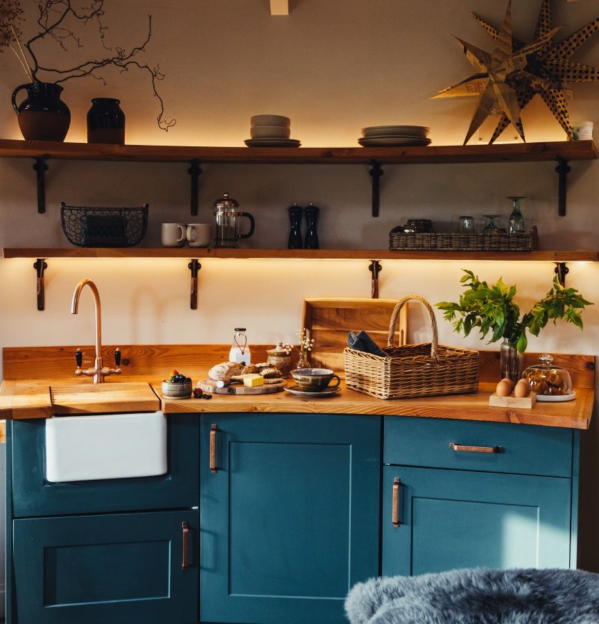 A cosy kitchen retreat featuring teal cabinets, wooden countertops, open shelves, a wicker basket, and decorative items at The Laundry Retreat Roundhouses