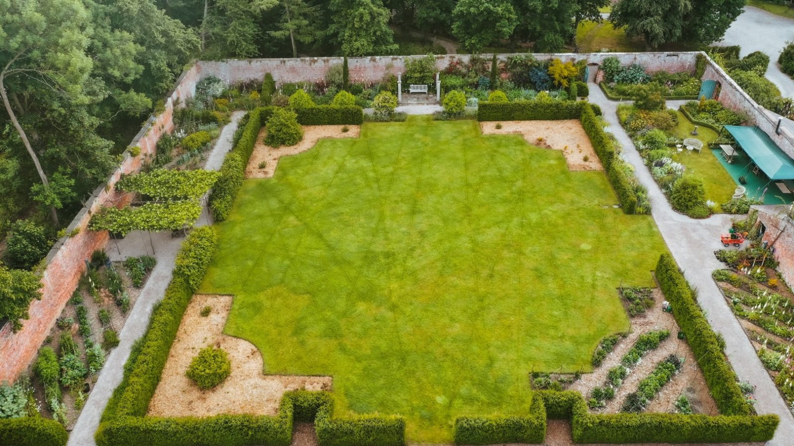 Aerial view of a serene retreat featuring a formal garden with a central lawn, surrounded by hedges, pathways, and various plants along the perimeter walls.