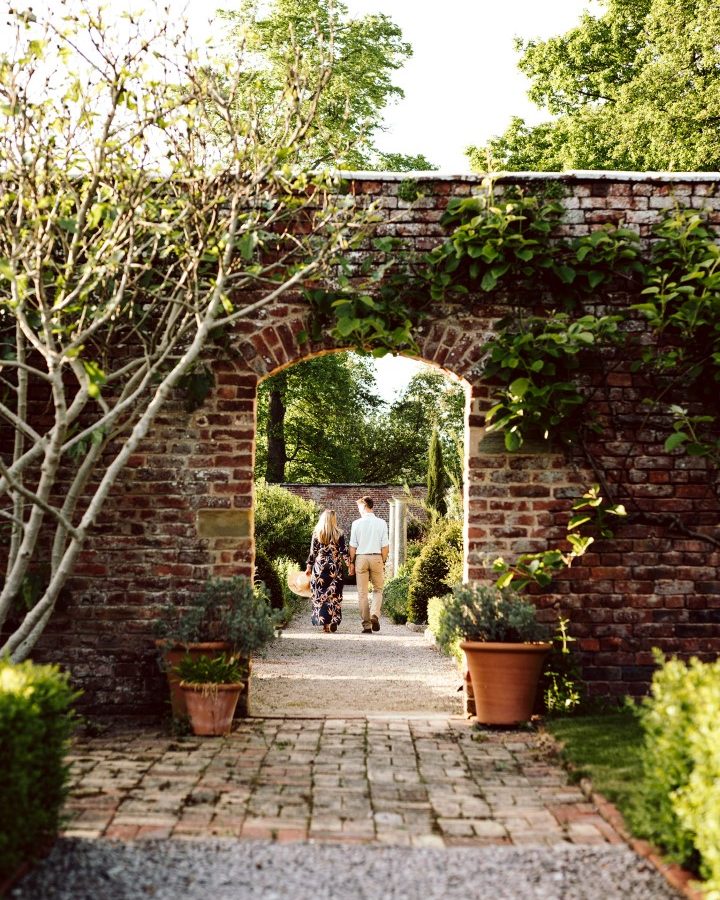 A couple walks through a brick archway in the garden of their charming hotel, surrounded by potted plants and trimmed hedges, on a sunny day.