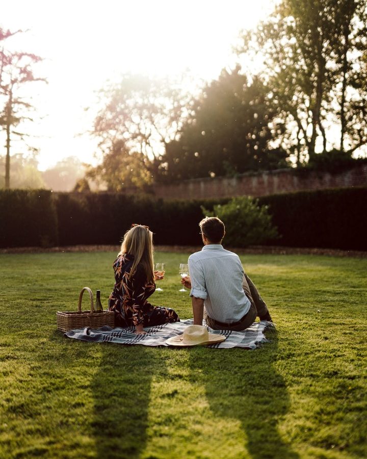 Two people sitting on a blanket in a sunny park, enjoying a peaceful retreat with drinks and a picnic basket nearby.