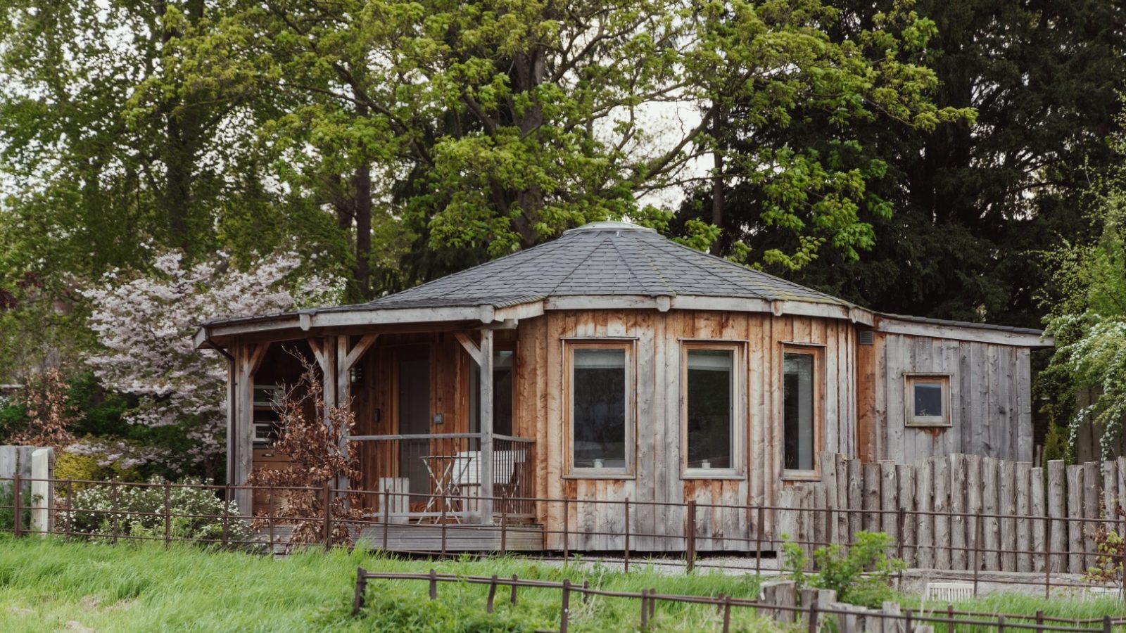 A rustic octagonal wooden cabin serves as the perfect retreat, with a cozy porch wrapped in trees and a wooden fence, nestled against a grassy foreground.