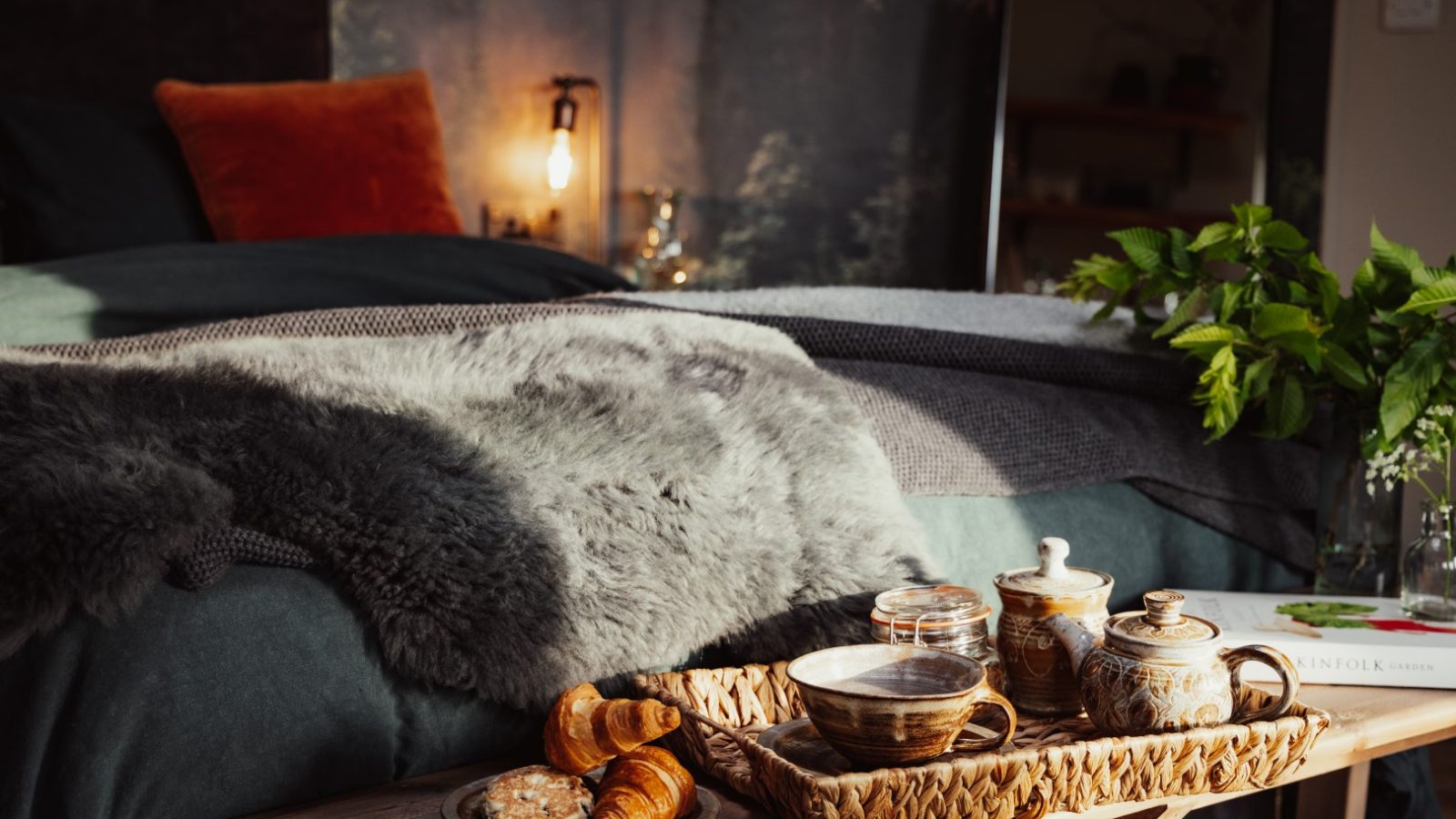 Cozy bedroom at The Laundry Retreat featuring a tray of tea, croissants, and cookies on a wooden bench. A fur blanket graces the bed, with lush plants in the background for a serene escape.