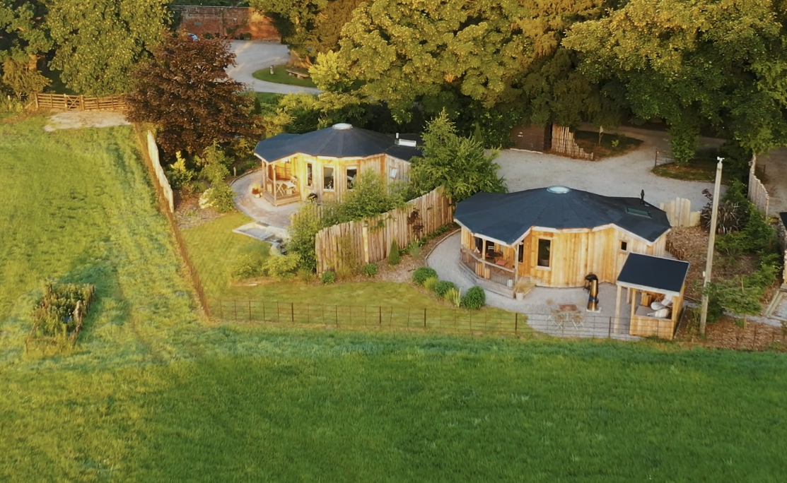 Aerial view of The Laundry Retreat, featuring two round wooden cabins with dark roofs nestled in a grassy area surrounded by trees and a nearby path.