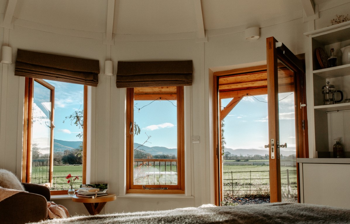 Interior view of a cozy retreat with open windows and door, overlooking a scenic landscape of mountains and fields under a blue sky.