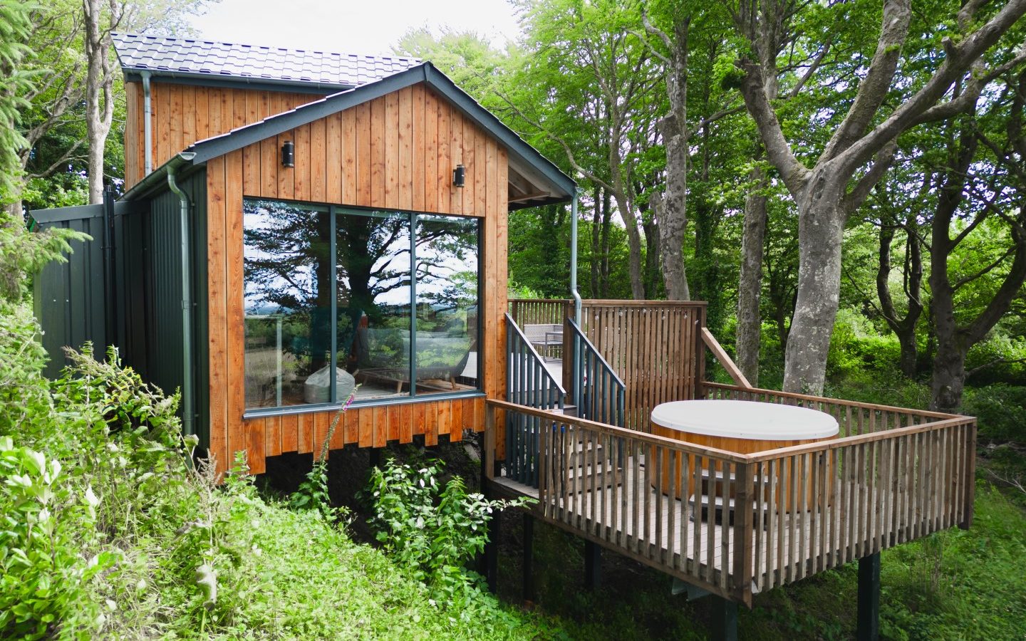 Cuckoos Hideaway, a wooden cabin with large glass windows and a deck featuring a hot tub, is surrounded by lush green trees.