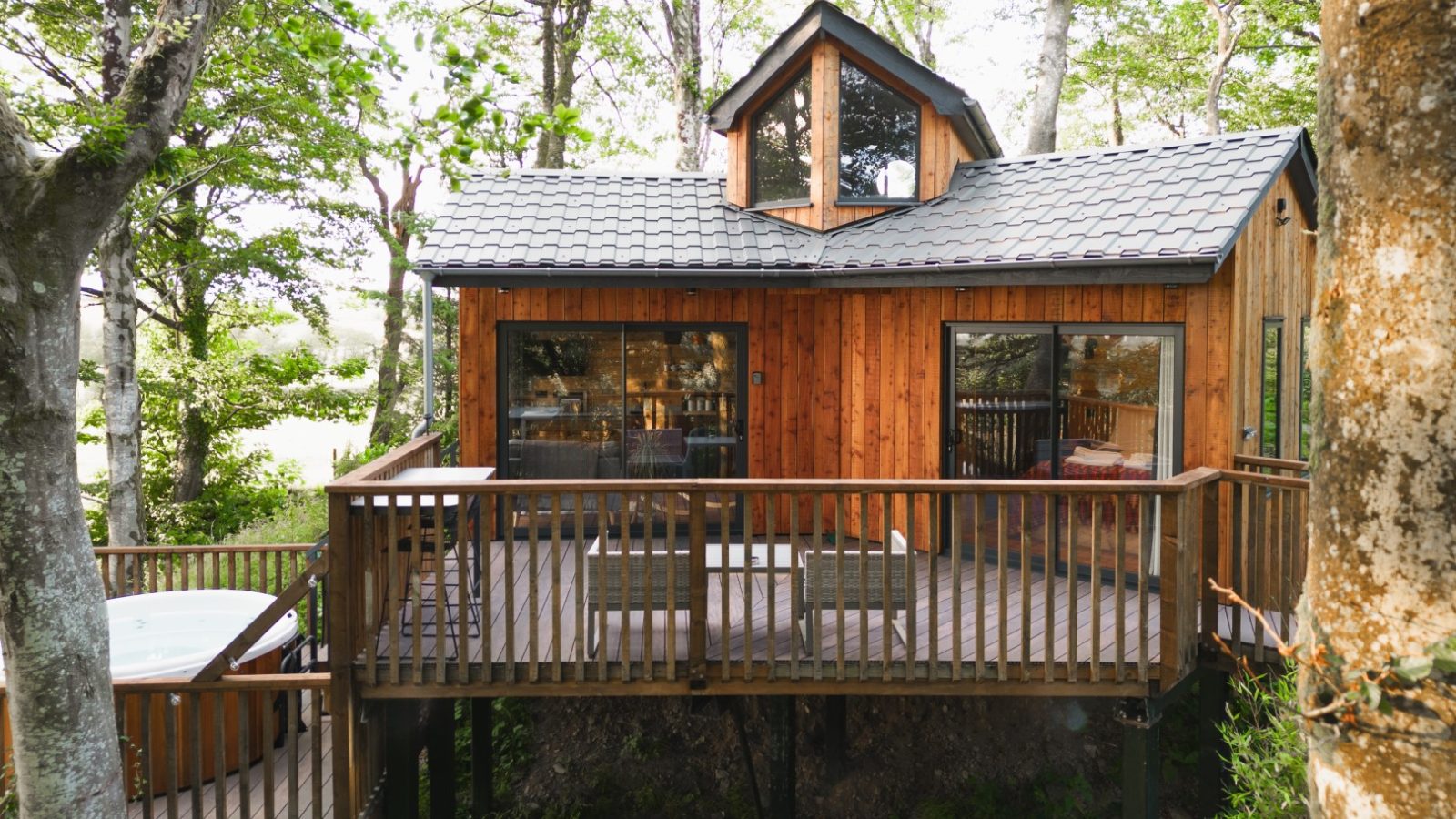 Cuckoos Hideaway is a wooden treehouse with large windows, a wraparound deck, and an outdoor hot tub, all nestled among lush green trees.