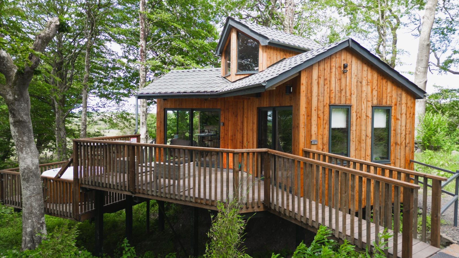 Cuckoos Hideaway is a small wooden cabin with large windows, a loft, and a wraparound deck, nestled among lush green trees.