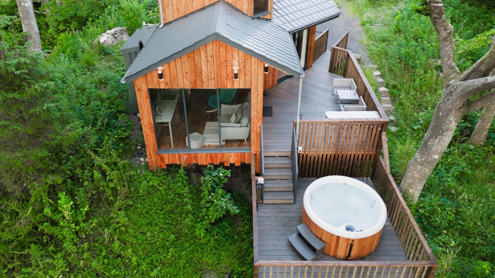 Aerial view of Cuckoos Hideaway, a wooden cabin with large windows, nestled among trees. It boasts a deck with a hot tub and seating area, perfect for relaxation.