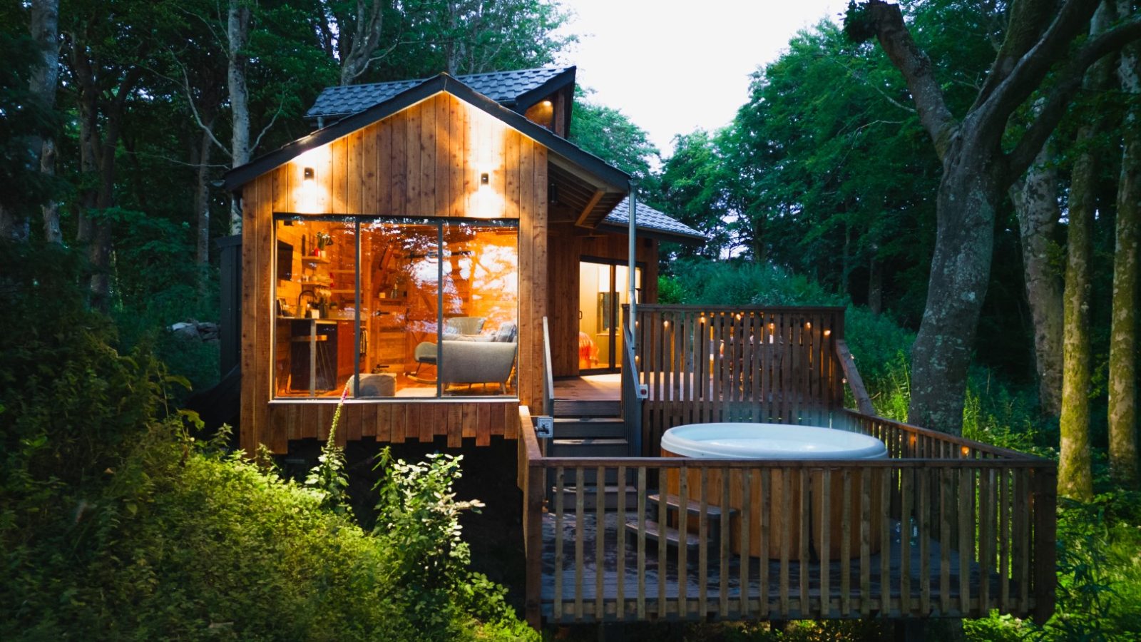 Cuckoos Hideaway is a cozy wooden cabin with large windows and an outdoor deck featuring a hot tub, nestled among trees in a serene forest setting.