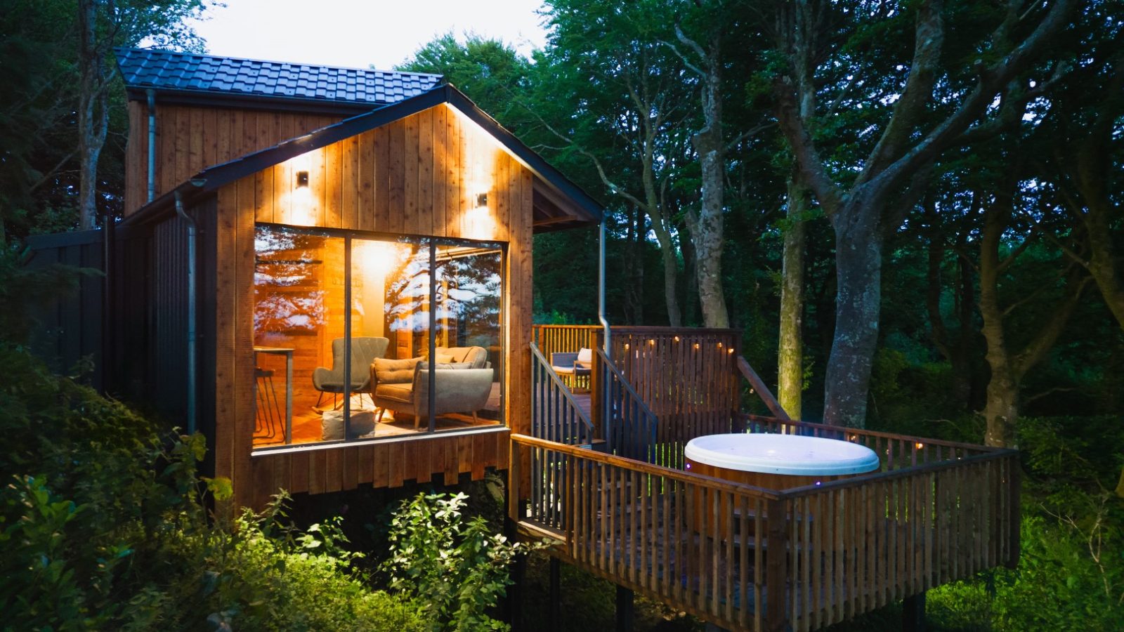 Escape to Cuckoos Hideaway, a modern wooden cabin with a large glass window and a deck featuring a hot tub. Surrounded by trees and warmly lit at dusk, it's the perfect retreat.