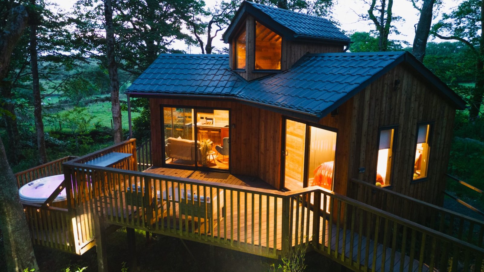 Cuckoos Hideaway, a cozy wooden cabin with a deck and large windows, nestles among the trees. Warm lights glow inside, inviting you to relax in the hot tub on the deck beneath a canopy of stars.