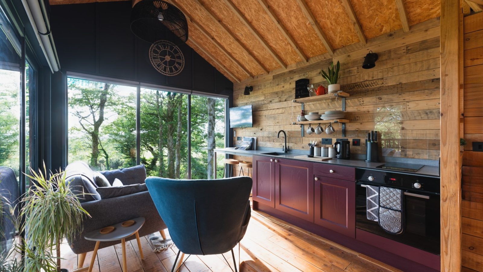 Welcome to Cuckoos Hideaway, a cozy cabin interior featuring a inviting kitchenette and seating area. The charming wooden walls and large windows frame breathtaking views of the surrounding forested area, creating the perfect serene retreat.