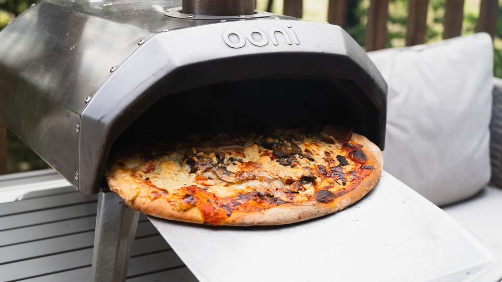 A pizza is being taken out of an Ooni outdoor pizza oven on a metallic pizza peel at Cuckoos Hideaway, set beautifully on an outdoor table.