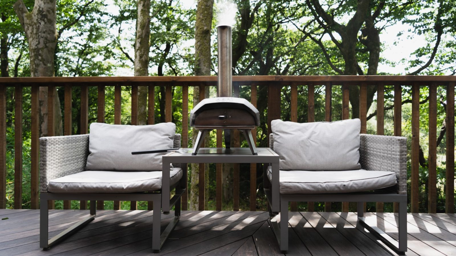 Nestled in Cuckoos Hideaway, two outdoor chairs with cushions flank a small table that cradles a wood-fired pizza oven, all set on a deck embraced by towering trees.