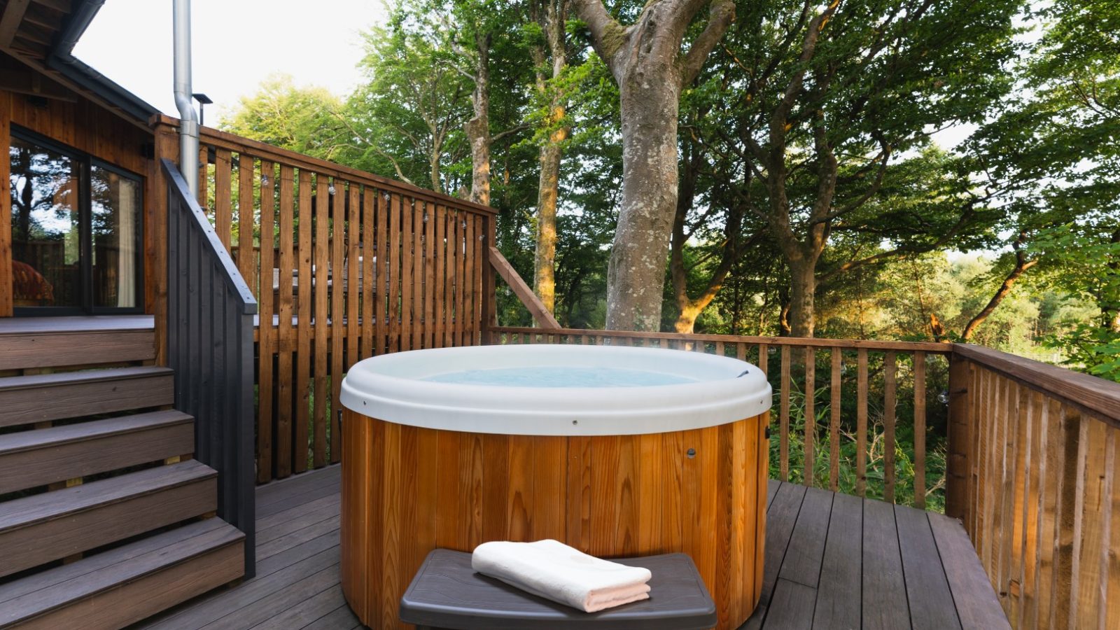 Nestled in Cuckoos Hideaway, a wooden hot tub sits on a deck embraced by trees, with folded towels neatly placed on a small table beside it.