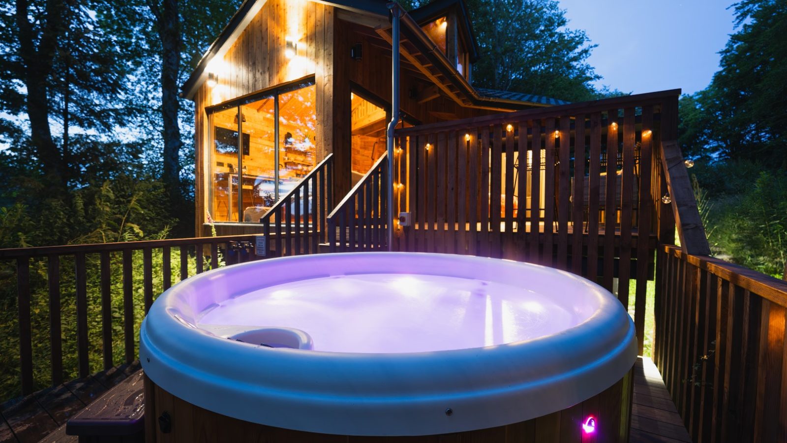 Cuckoos Hideaway: A wooden cabin with large windows surrounded by trees, featuring a lit hot tub on a deck, photographed during twilight.