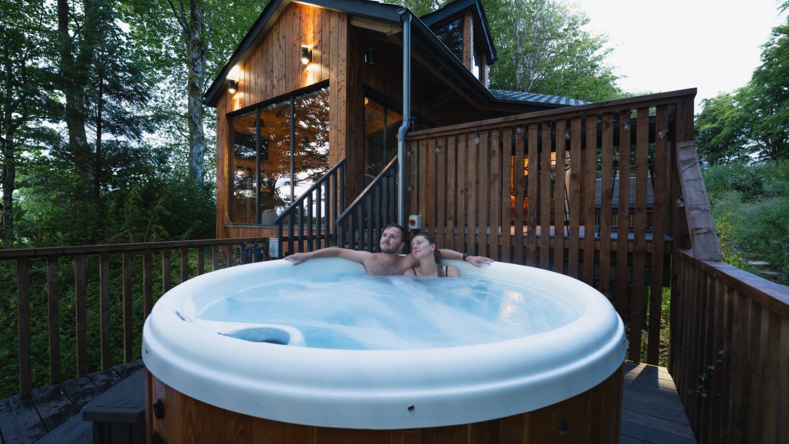 At Cuckoos Hideaway, a couple unwinds in an outdoor hot tub on a wooden deck, set against the backdrop of rustic cabin charm and towering trees.