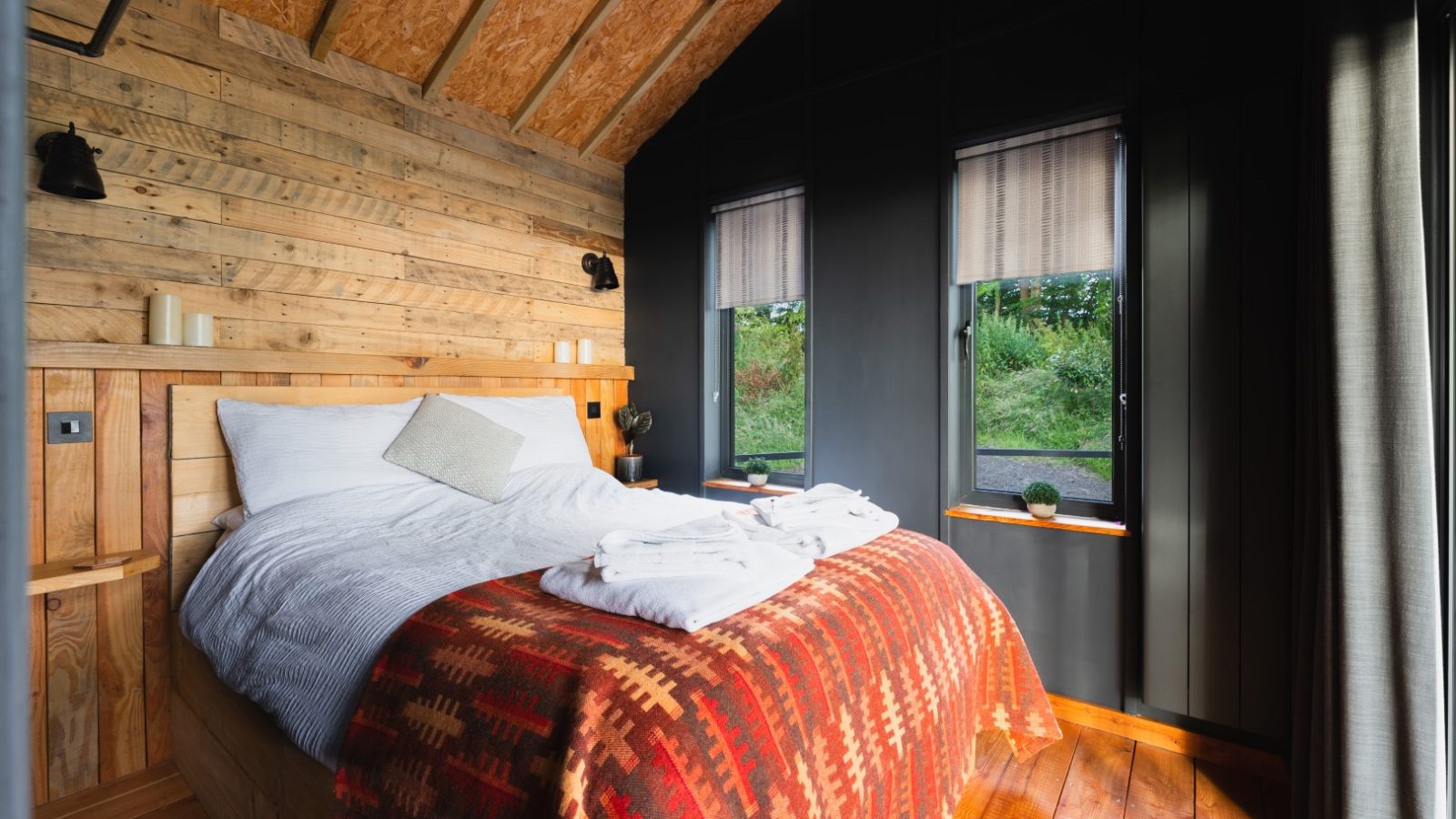 Cozy bedroom in Cuckoos Hideaway with a bed, wooden walls, and two windows. The bed features a patterned red blanket and neatly folded white towels.