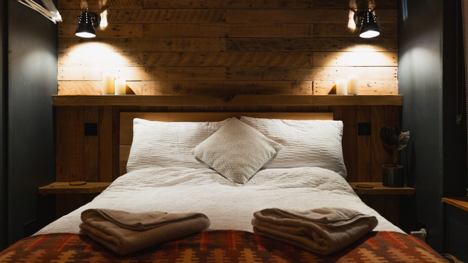 Cozy bedroom at Cuckoos Hideaway with a wooden headboard, white bedding, folded blankets at the end, and small wall-mounted lamps above.