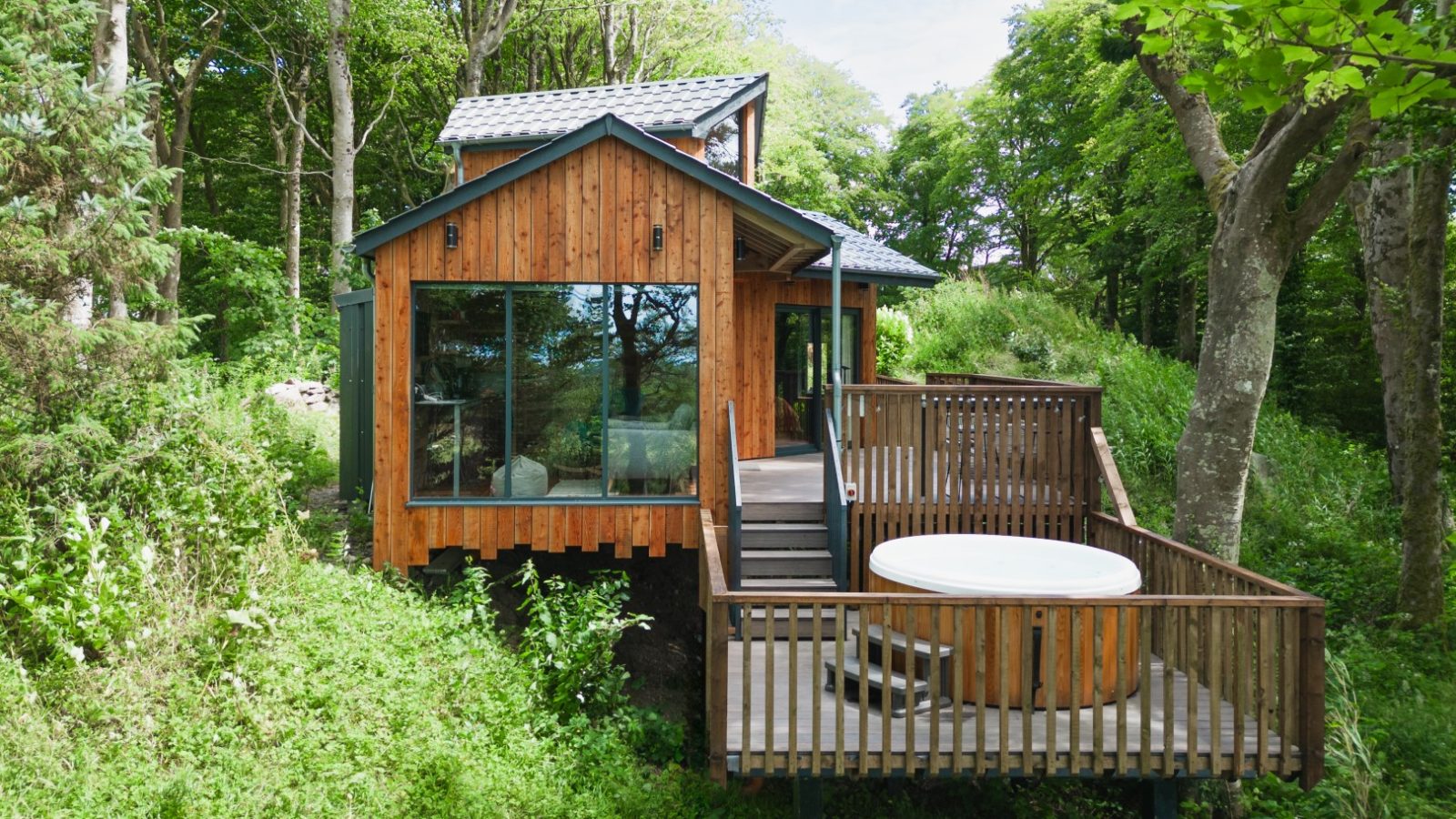 Welcome to Cuckoos Hideaway, a charming wooden cabin with large windows and a hot tub on a deck, surrounded by trees and lush greenery.