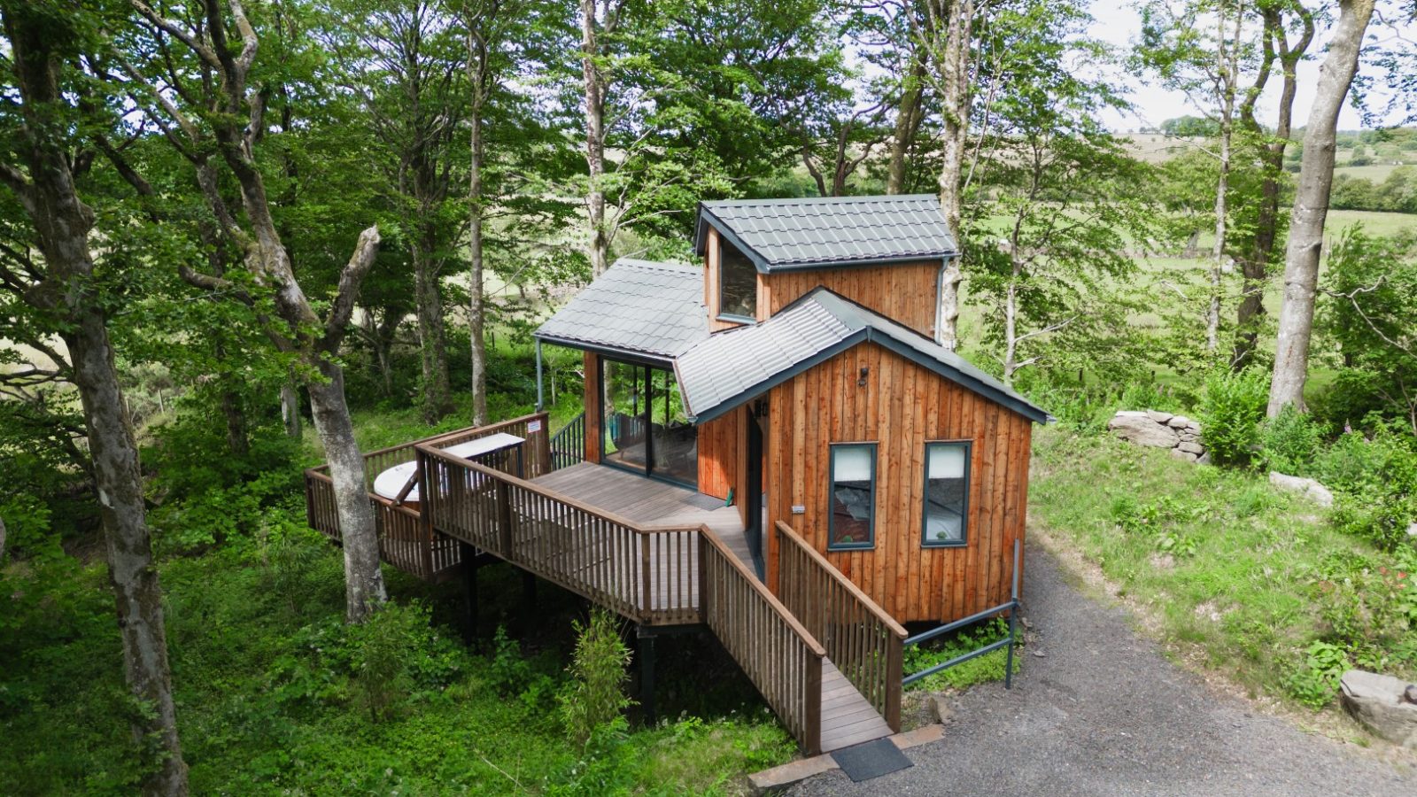 Nestled among the trees, Cuckoos Hideaway is a small wooden cabin featuring a wraparound deck and a gravel path leading to its charming entrance.