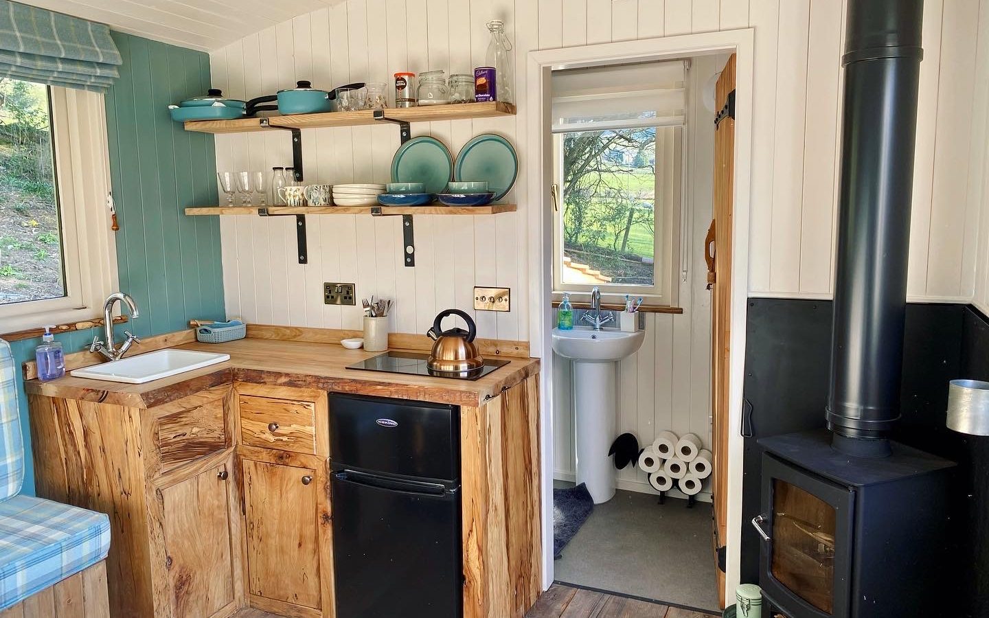 Cozy wooden kitchen reminiscent of Caban Eric, with a small sink, mini fridge, and shelves holding dishes. A kettle sits on the counter, and through an open door, a compact bathroom is visible. To the right, a wood-burning stove with a chimney stands. A scenic outdoor view unfolds through the window.