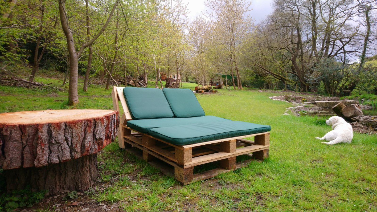 A wooden pallet sofa with green cushions rests on lush grass near a tree stump table in Caban Eric's serene garden. A white dog relaxes nearby, enveloped by trees and a cloudy sky, creating a tranquil haven.