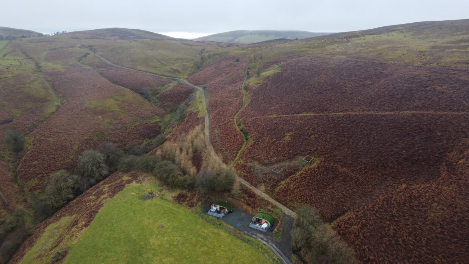 An aerial view reveals a narrow road winding through hilly terrain, adorned with patches of grass. Nestled close to the road are two small buildings, offering scenic accommodations at Beacon View Pods, the perfect glamping escape.