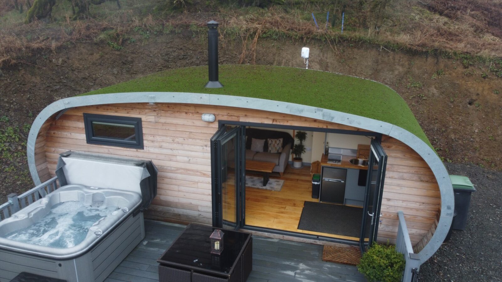 A small wooden cabin with a green roof, open doors showing the interior, stands beside a deck with a hot tub and seating area, nestled among the scenic charm of Beacon View Pods.