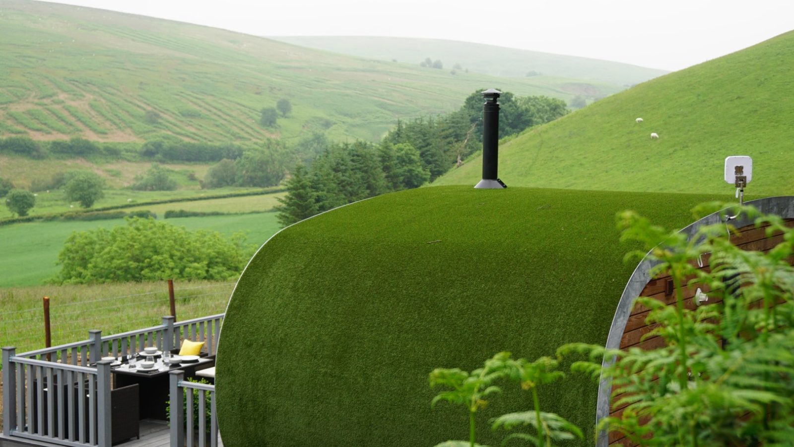 A small, rounded green-roofed pod with a chimney sits on a wooden deck in a lush, hilly landscape. The deck has a set table with chairs, offering an incredible view of rolling green hills under a misty sky. This is the serene experience at Beacon View Pods.