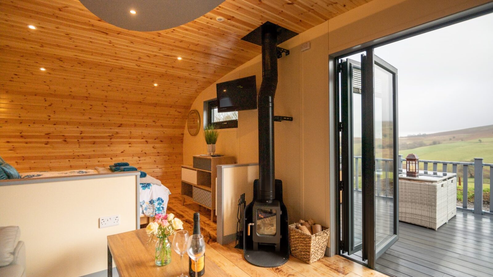 The cozy wooden interior of this pod features a bed, table, and stove, with open doors leading to a deck that offers stunning Beacon View countryside vistas.