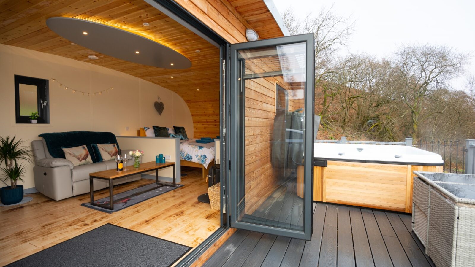 The interior view of this cozy wooden cabin, aptly named Beacon View, features an open glass door revealing a snug sofa, a rustic table and bed, and leads to an outdoor hot tub on the deck—a perfect retreat nestled among nature’s pods.