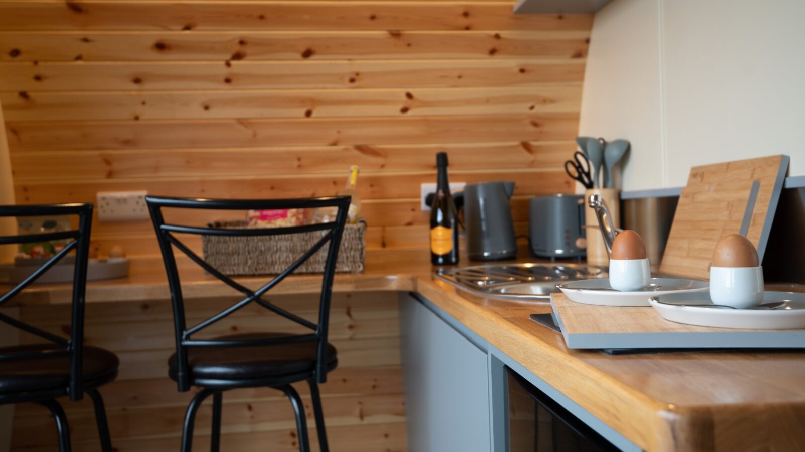 Cozy kitchen with wooden walls, two stools, countertop appliances, and soft-boiled eggs on plates with egg cups, offering a glimpse of Beacon View Pods.