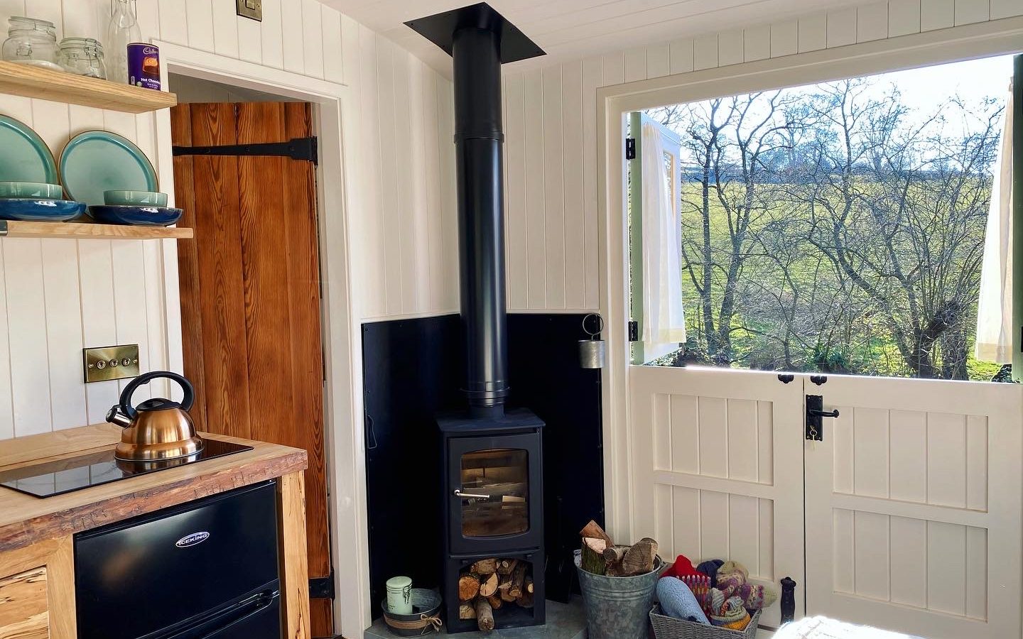 Cozy room at Caban Eric with a wood-burning stove, a basket of logs, and an open Dutch door revealing sunny views of trees and fields. Shelves with blue-green dishes, a wooden cabinet, and a kettle on the quaint stove add warmth to the rustic setting.