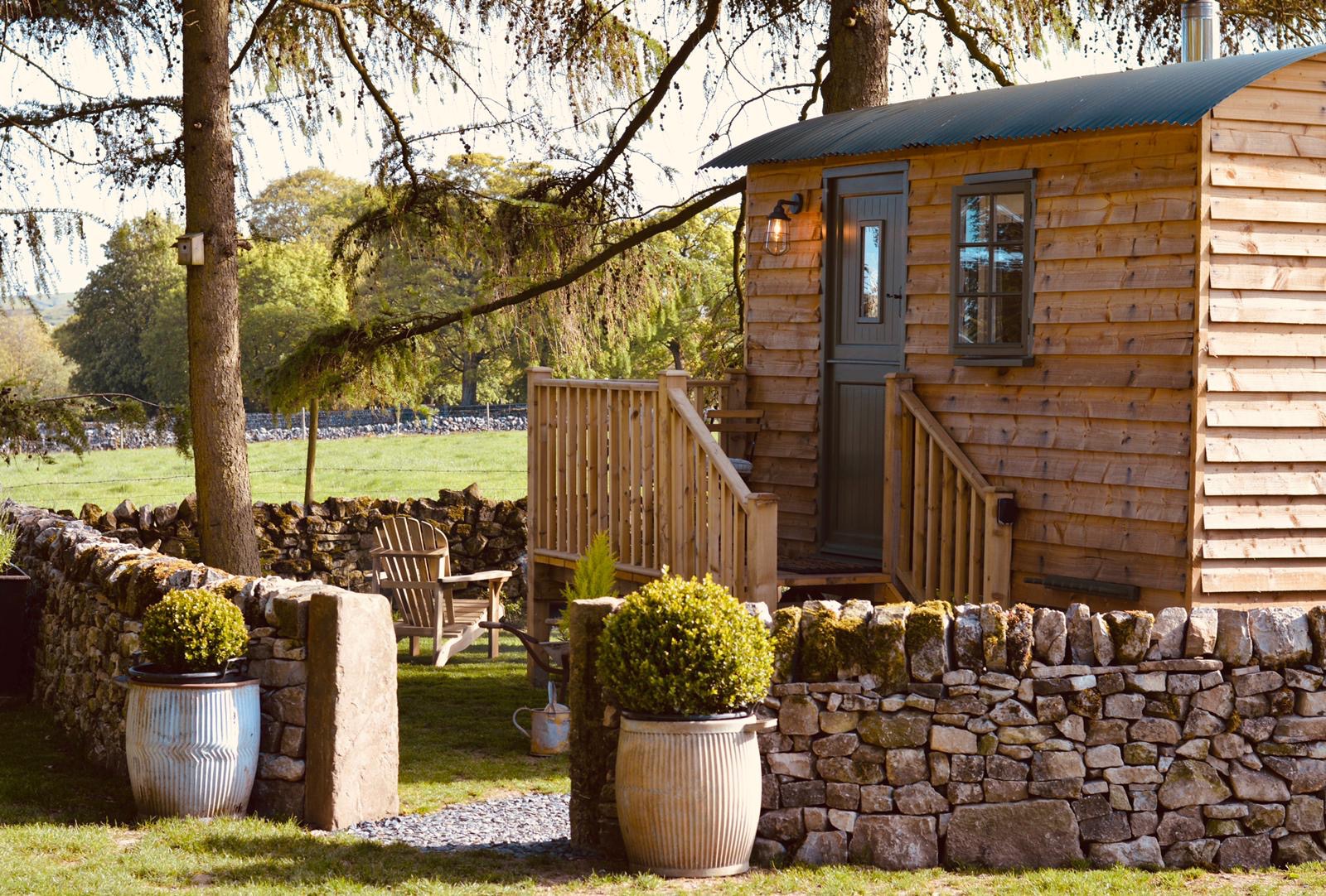 A wooden tiny house, perfect for glamping accommodation, features steps and a small deck. It's surrounded by stone walls, lush trees, and potted plants in a tranquil grassy area.