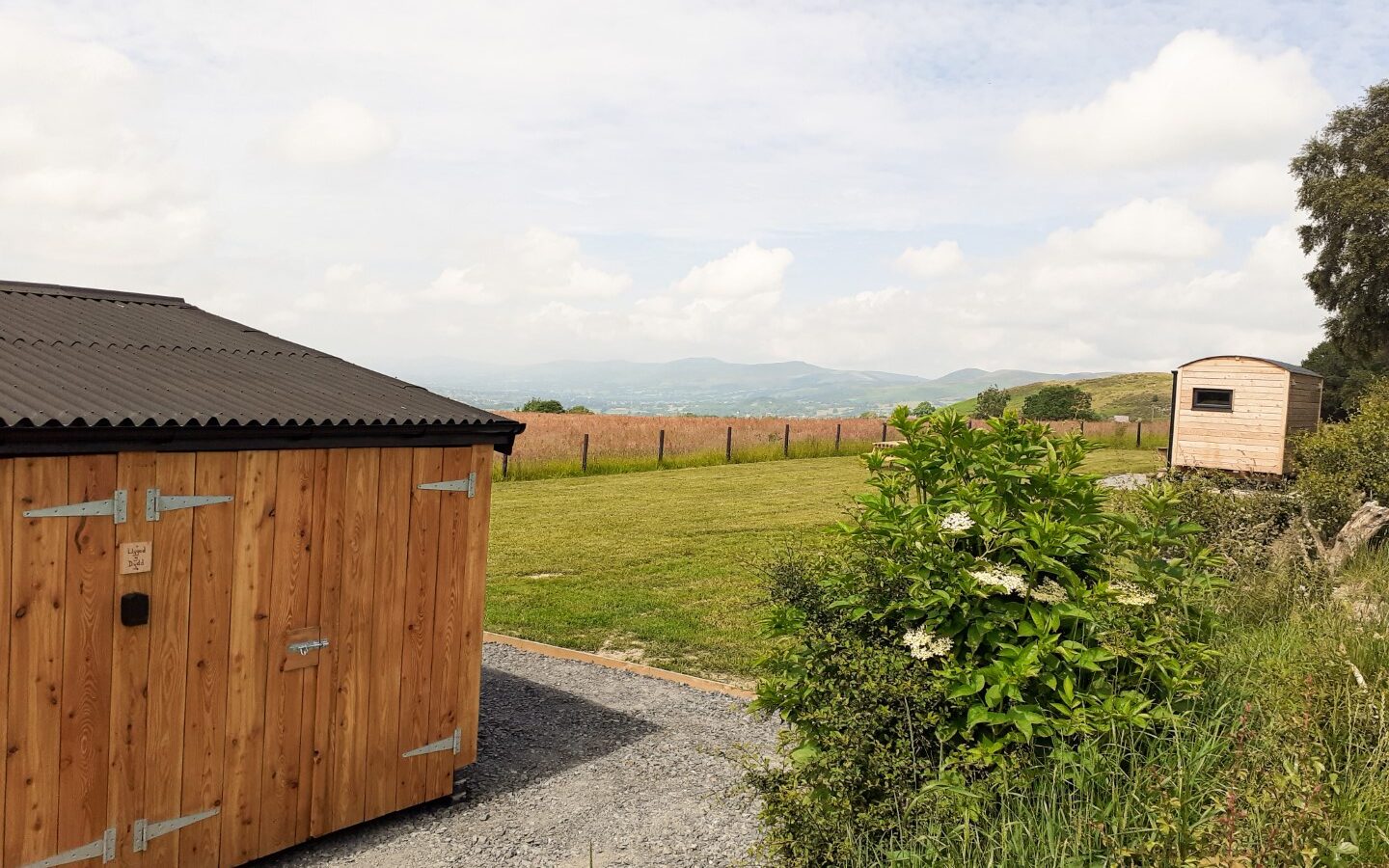 On the grassy landscape of Cae Main, wooden sheds stand serenely, their rustic charm mirrored by the rolling hills and a cloudy sky. These quaint structures evoke the essence of a shepherd's hut nestled in picturesque tranquility.