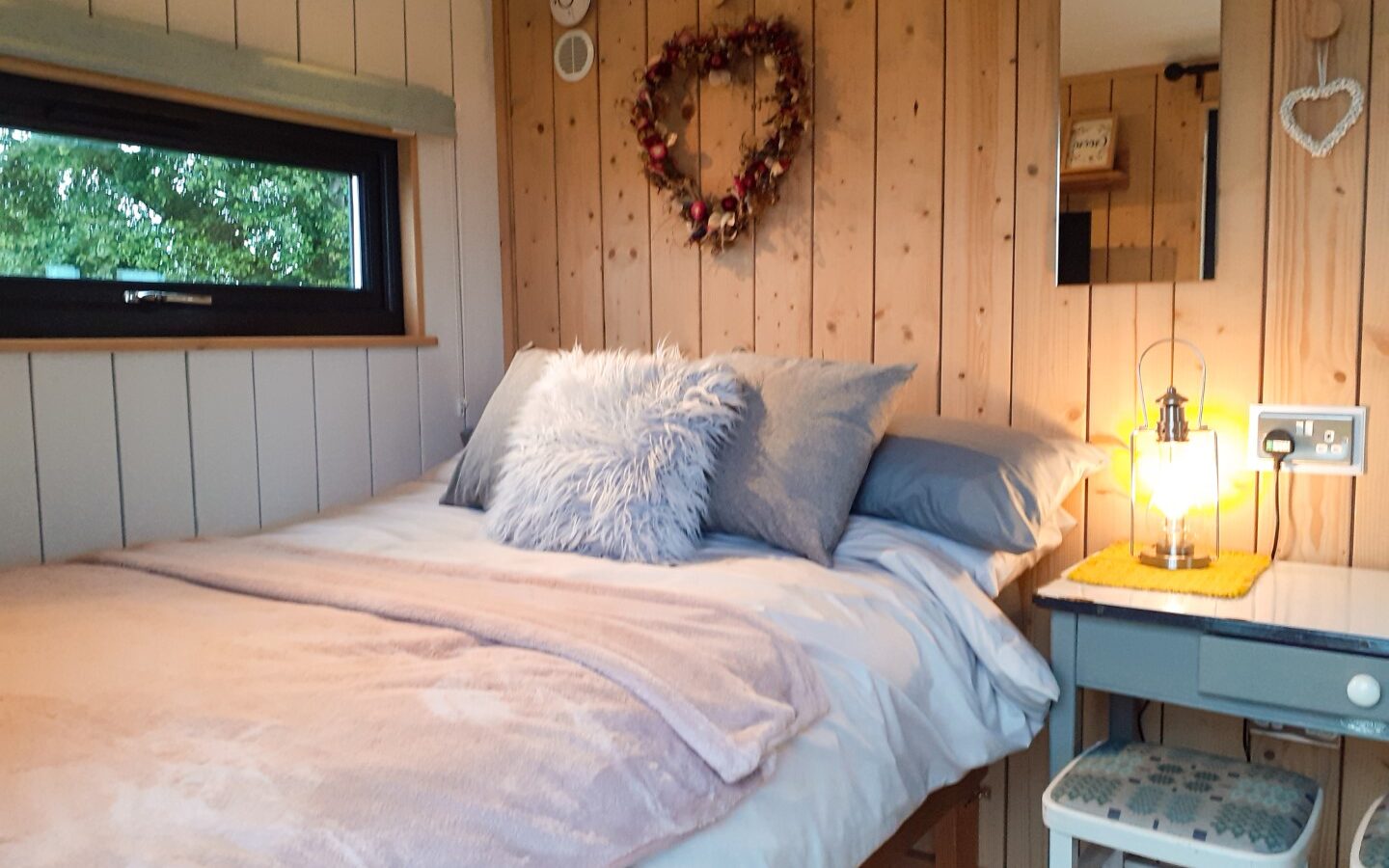 This cozy Shepherds Hut features a wooden accent wall, a snug double bed with pillows and blankets, and a small bedside table with a lamp and charming decor, making it the perfect retreat at Cae Main.