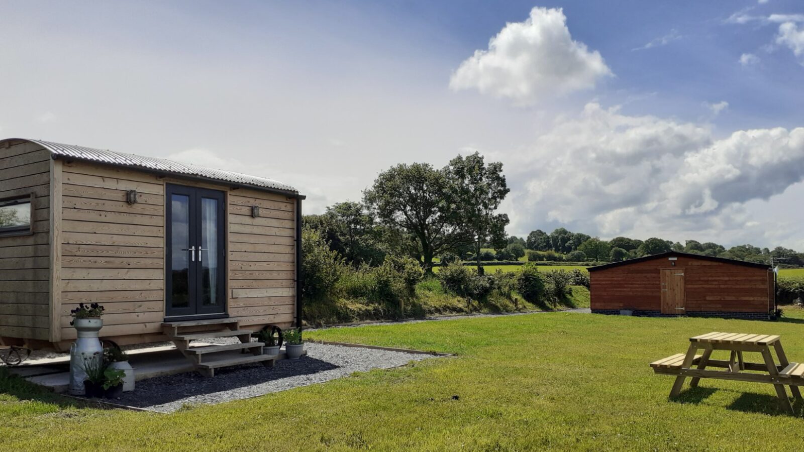 A charming shepherd's hut sits on the grass with a bench nearby, offering unique accommodation. In the background, Cae Main's larger building stands under a partly cloudy sky.