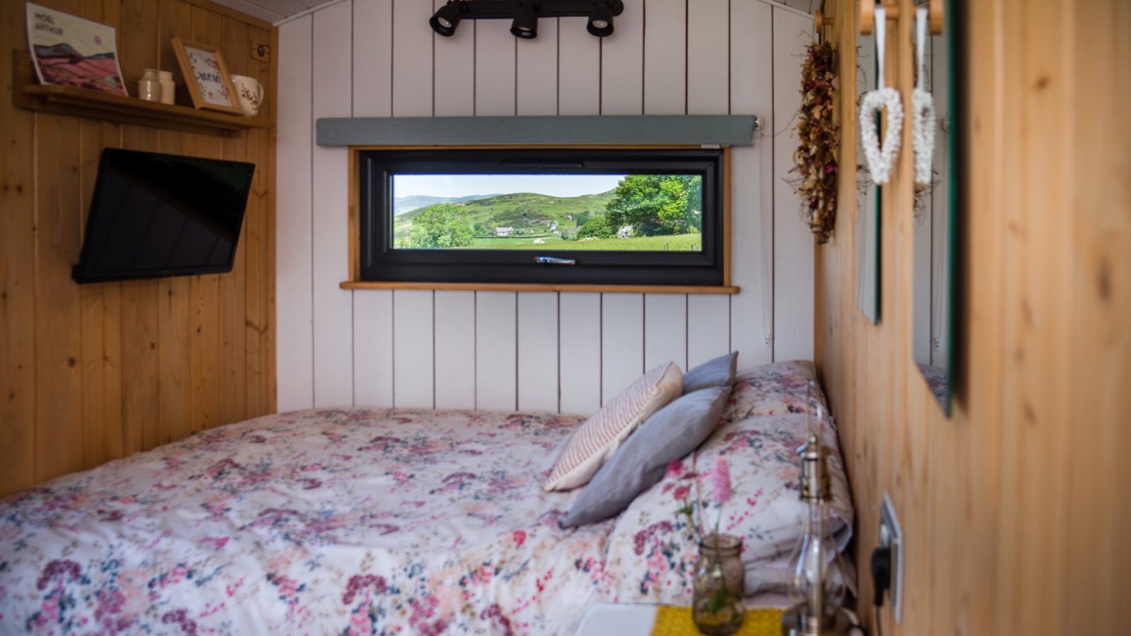 Cozy Cae Main cabin interior with floral bedspread, green hill views, wall art, and a small shelf with books.