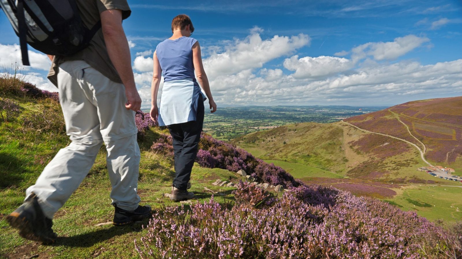 Two people hiking down a scenic hillside near Cae Main, surrounded by heather, overlook the countryside under a partly cloudy sky.